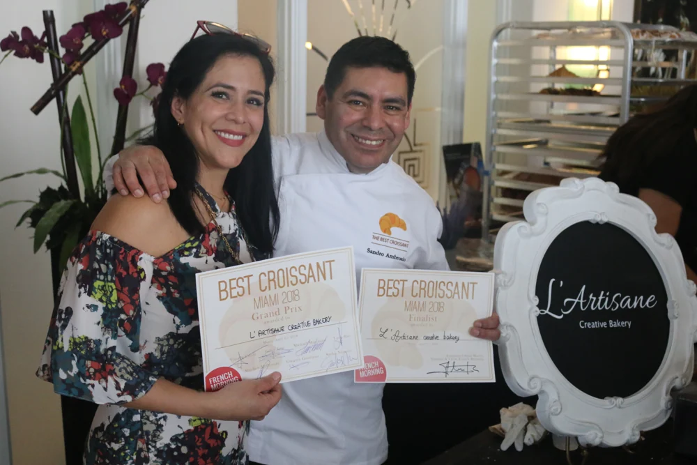 Two members of the L’Artisane team celebrating with certificates beside the bakery’s original logo